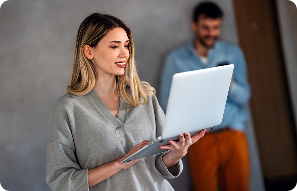 Mujer sosteniendo y mirando un computador, al fondo se ve un hombre mirando su celular.