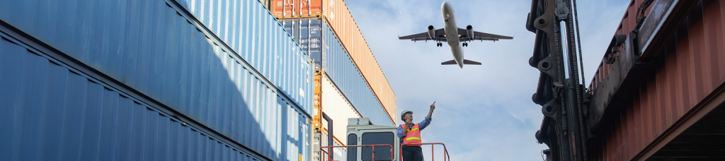 Vista de un hombre de pie señalando un avión que está volando. Al lado izquierdo hay contenedores.